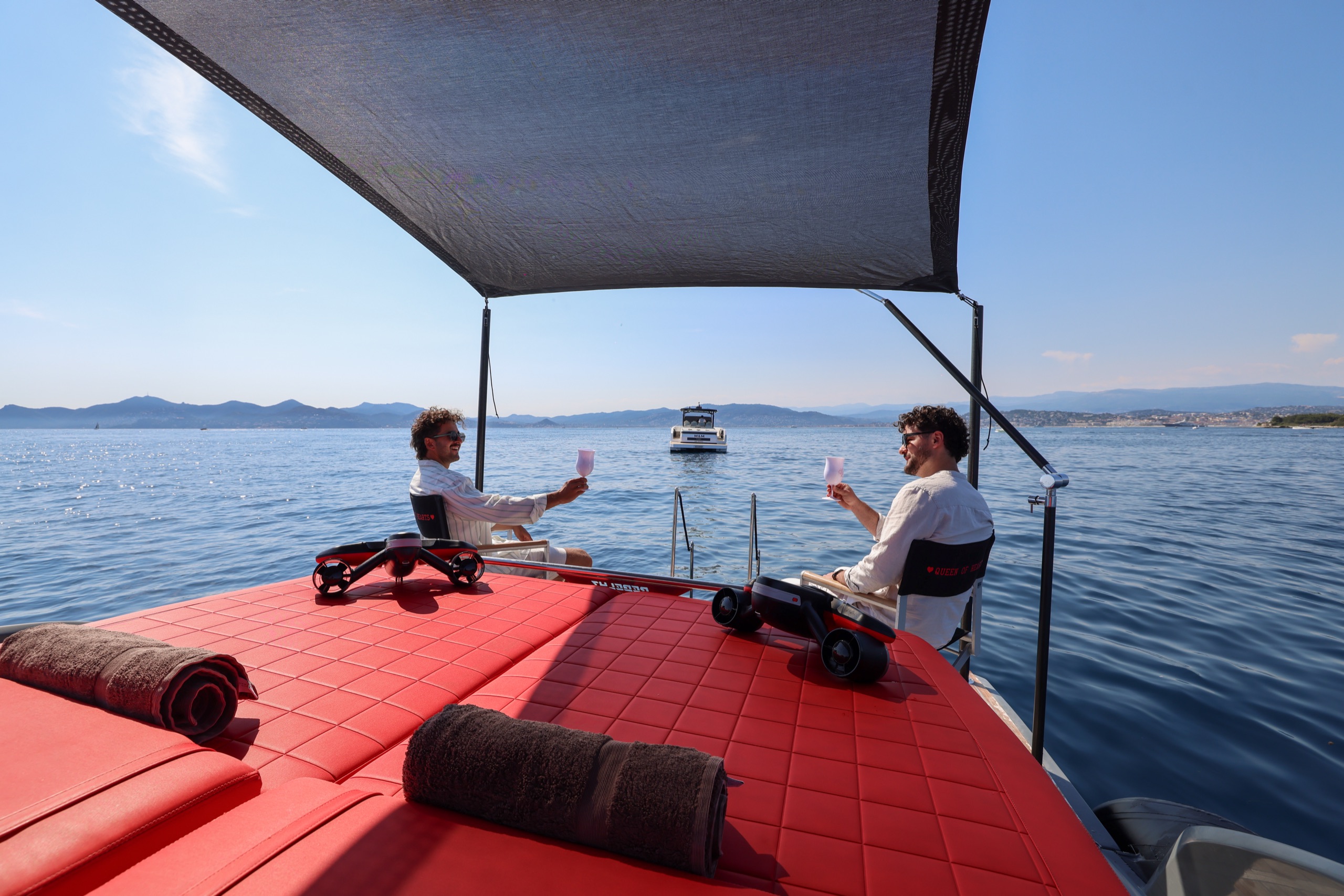 Guests raising a toast under the shade sail on Queen of Hearts — red sunbeds, seabobs ready, the coast behind