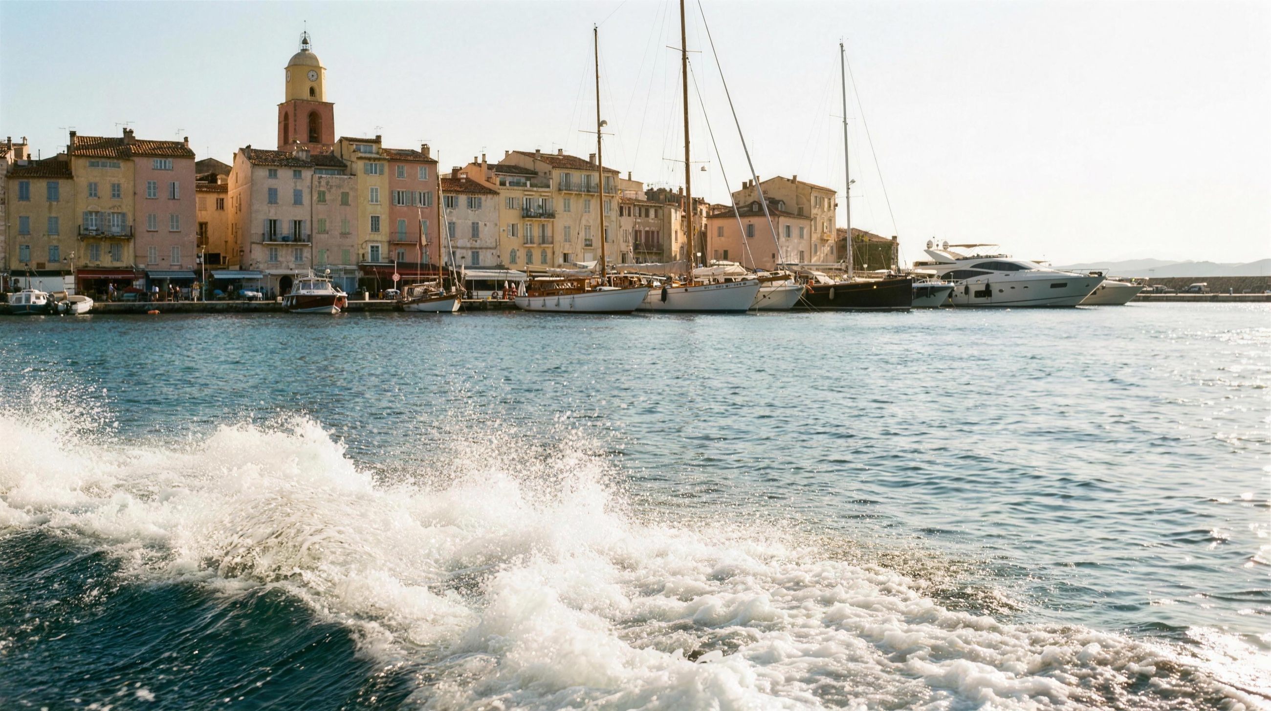 Wake spray on the water arriving at Saint-Tropez old port — the ochre facades, bell tower and moored yachts beyond